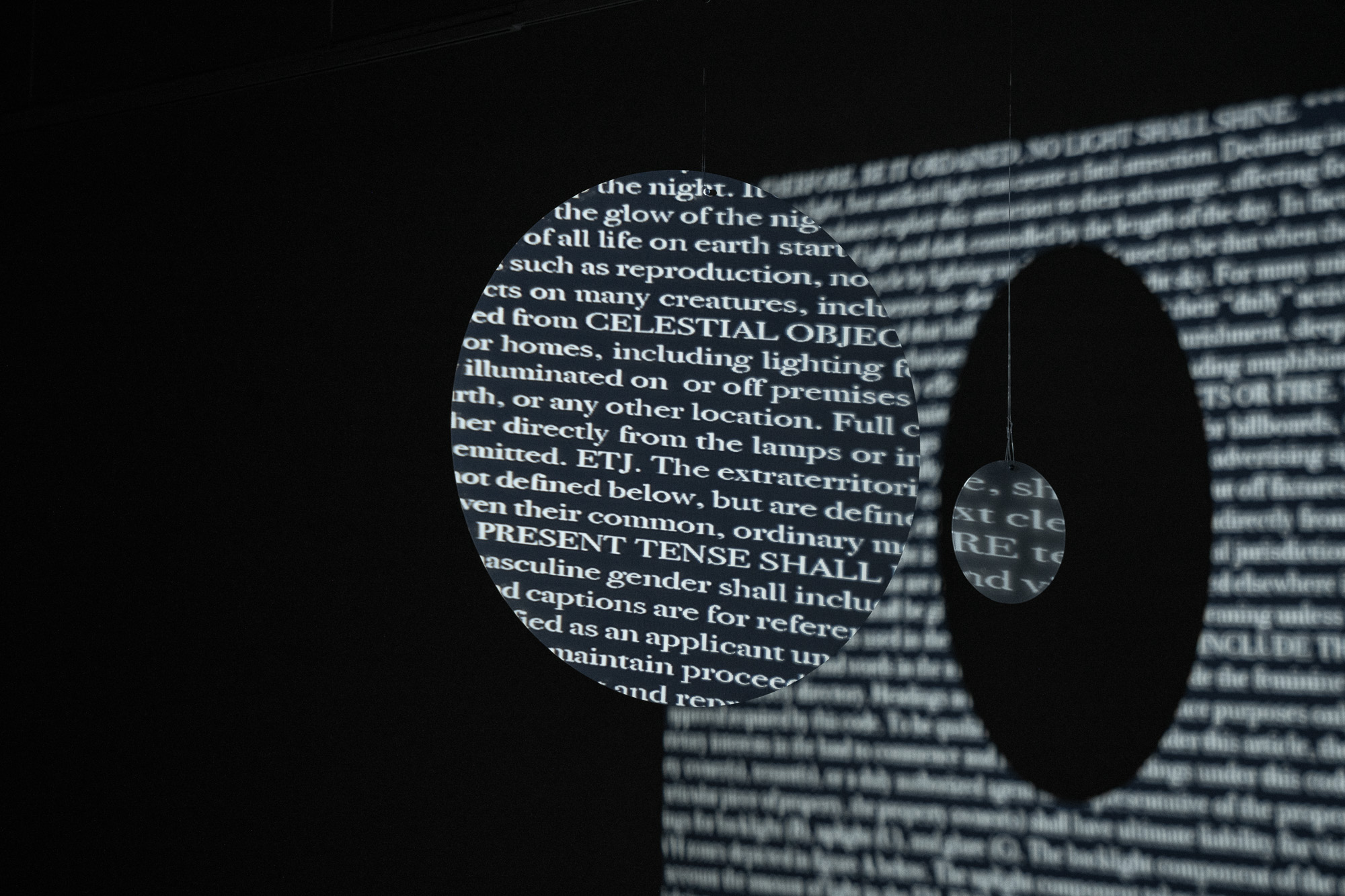 Chad Ress, multimedia art of writing with words in white on black background contained in a floating circle, next to white writing on a dark wall, the shadow of the circle visible.