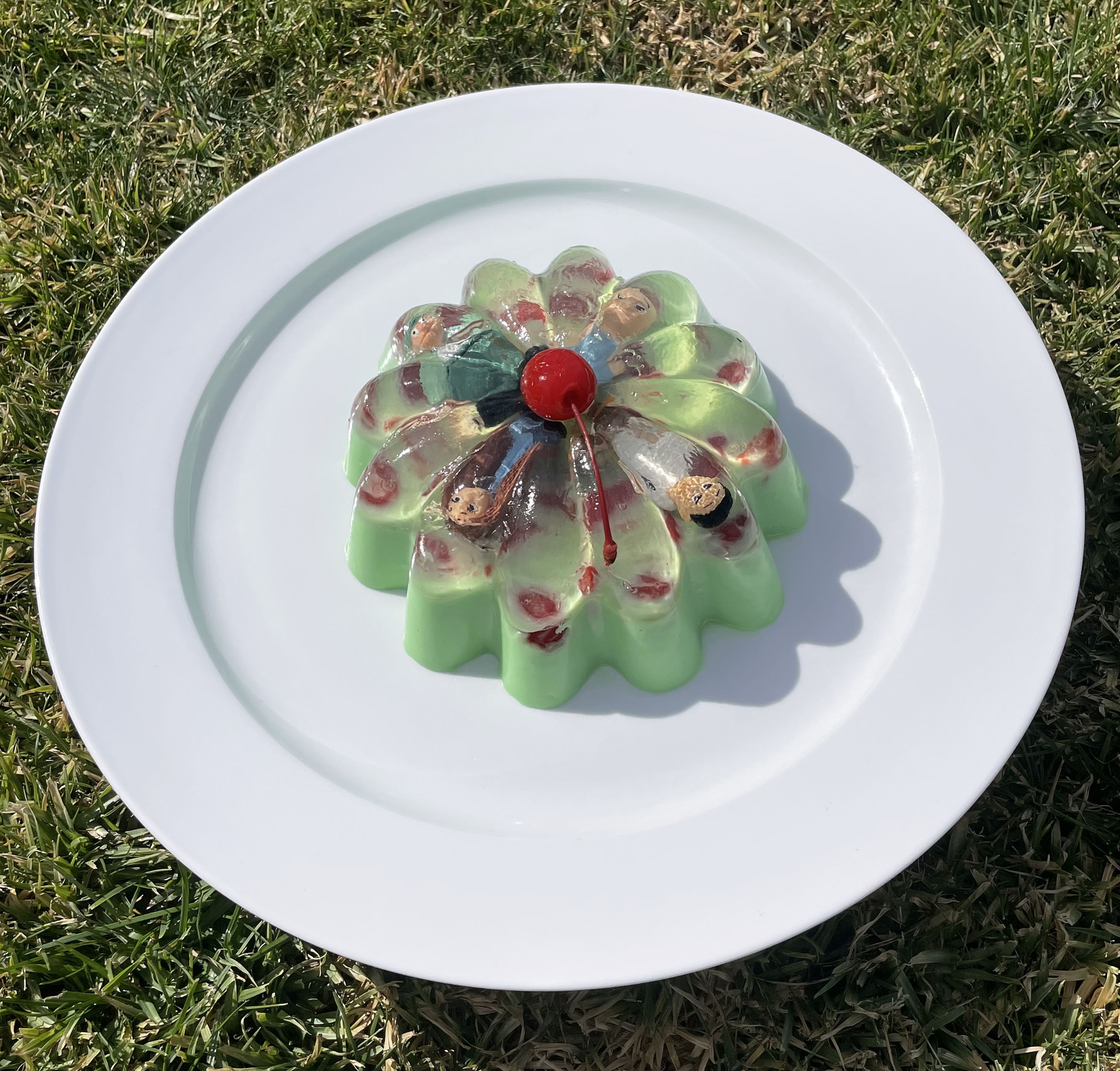 Marissa de la Peña art, green jello mold with four cartoon-like figures encased inside, with a cherry on top. Plated on a white plate with plate resting on green grass.